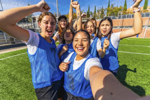 Group of excited multiracial women soccer players in blue vests and white t shirts celebrating victory on a green sports field, taking a joyful selfie on a sunny day