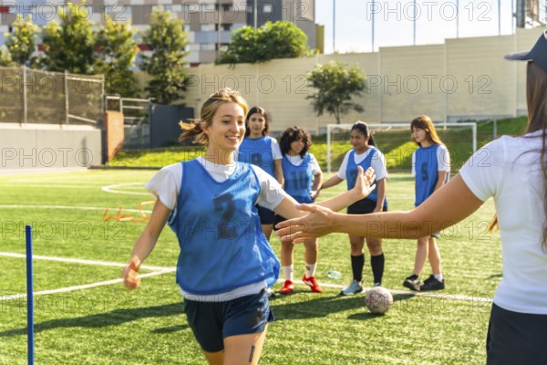 Young female soccer player in a blue bib high fiving her coach on a green artificial turf field, celebrating achievement and teamwork during training with teammates in the background