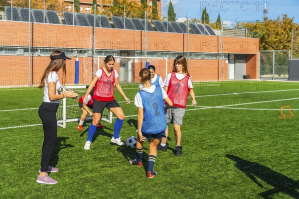 Girls soccer team receiving instructions from their female coach while practicing ball control and passing drills on an artificial turf field, developing teamwork and football skills