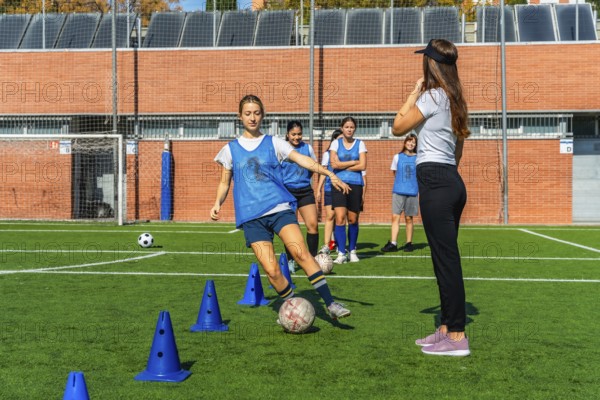 Group of diverse young women soccer players training with their coach on a green artificial turf field, practicing dribbling skills around blue cones during a sports session