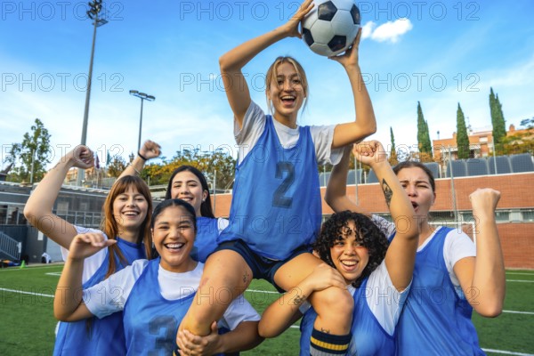 Group of diverse women soccer players cheering and lifting a teammate with a soccer ball on a green field, expressing happiness and teamwork after a competitive match