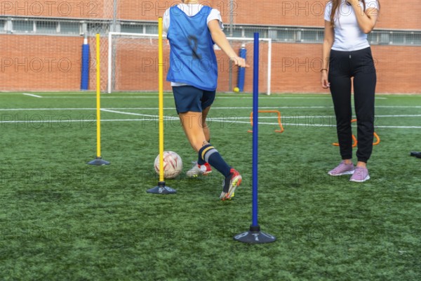 Young woman in a blue bib practicing soccer dribbling skills, maneuvering a ball through training poles on a vibrant green artificial turf field with a coach observing