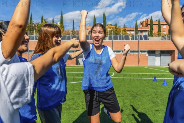 Excited female soccer players celebrating success and victory together, raising their arms in jubilation on a green artificial turf field after a game
