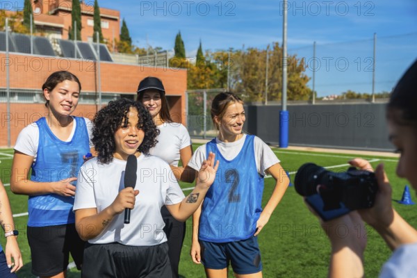 Female football player talking into a microphone, sharing her thoughts and insights about the team's performance, while other players stand behind her and a person films the interview