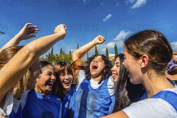 Young women soccer players cheering with arms raised in celebration, smiling and embracing after victory under a blue sky, showing teamwork, joy, friendship and empowerment