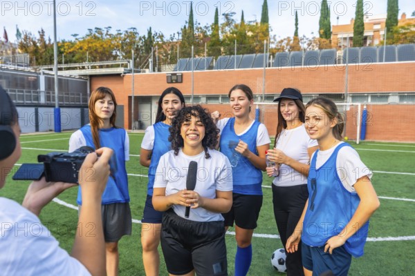 Female football players and their coach standing on a green field after a game, giving an interview to a reporter holding a microphone and being filmed by a videographer