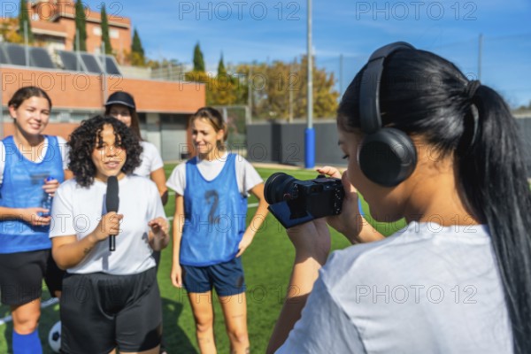 Videographer with headphones filming a reporter holding a microphone and interviewing a group of diverse young women soccer players on a green sports field