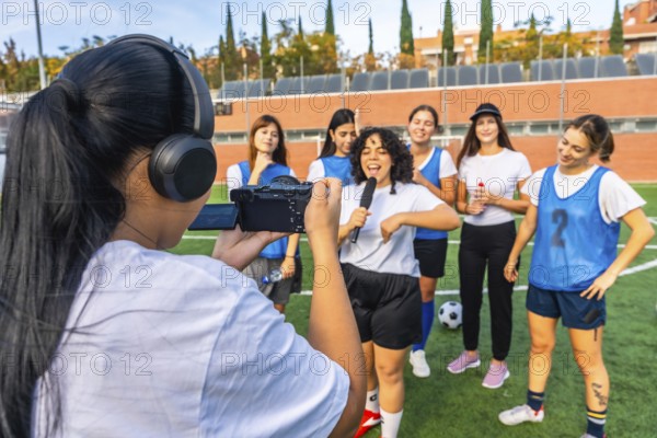 Woman videographer in headphones filming a dynamic interview with a joyful woman holding a microphone and her diverse women soccer teammates on a green sports field