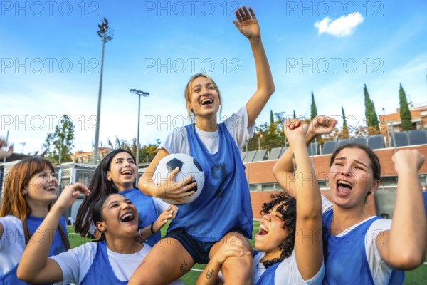 Joyful female soccer players lifting a teammate with the ball overhead, celebrating a win together on the green field under a clear blue sky, showing teamwork and success in sport