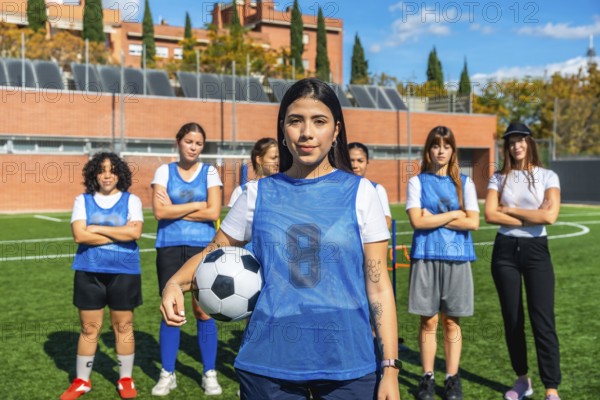 Young female soccer players standing on a sunny artificial turf field, with one player holding a football and looking at the camera, representing teamwork and active lifestyle