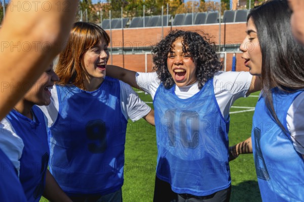 Young diverse women football players joining arms in a huddle, enthusiastically celebrating a victory or goal on a sunny outdoor sports pitch, showing unity, achievement and teamwork