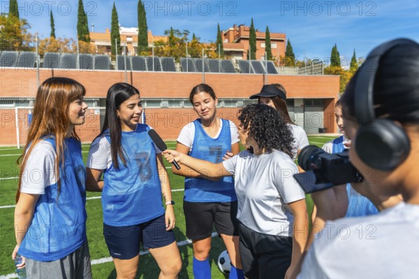 Women soccer team players standing on a green sports field, one player holding a microphone while interviewing two teammates, with a videographer filming the scene