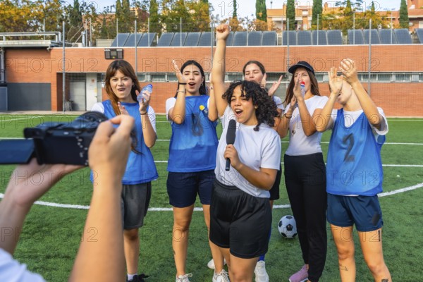 Women's football team standing on grass field, cheering and celebrating a win as one player holds a microphone, giving an interview to a person recording the event