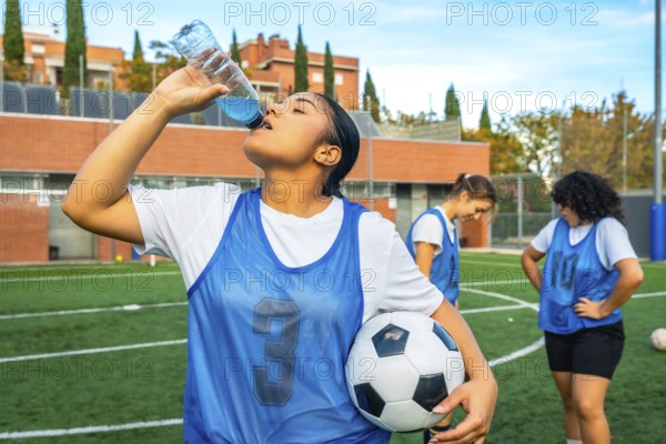 Young woman soccer player in a blue bib holding a football, refreshing herself by drinking a sports energy drink on a synthetic turf field after practice, with teammates in the background