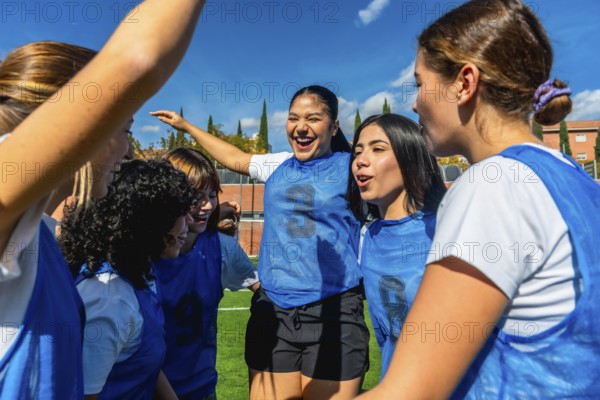 Group of diverse women's soccer players huddling on the field, actively celebrating their success and showing vibrant team spirit and strong teamwork under a bright blue sky