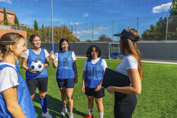 Women soccer players in blue pinnies listening attentively to their female coach explaining strategies and guiding the team on a sunny artificial grass field