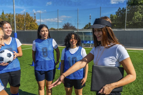 Coach, wearing a visor and holding a folder, explaining strategy to a smiling female soccer team on a green artificial turf field under a clear blue sky