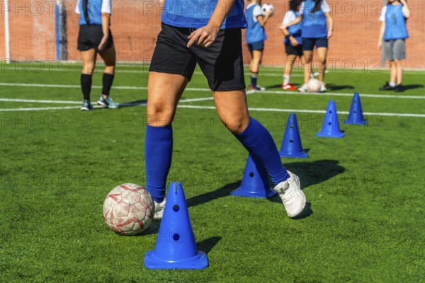 Female football player dribbling a soccer ball between blue training cones on a green artificial turf field, her teammates practicing in the background