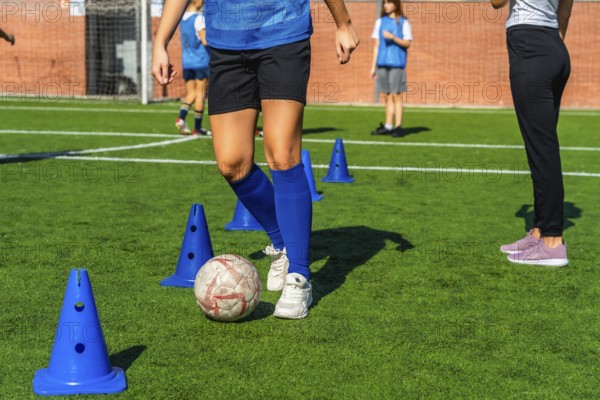 Young girl in a blue jersey practicing soccer dribbling skills around orange cones on a green artificial turf field, training for the sport with her team on a sunny day