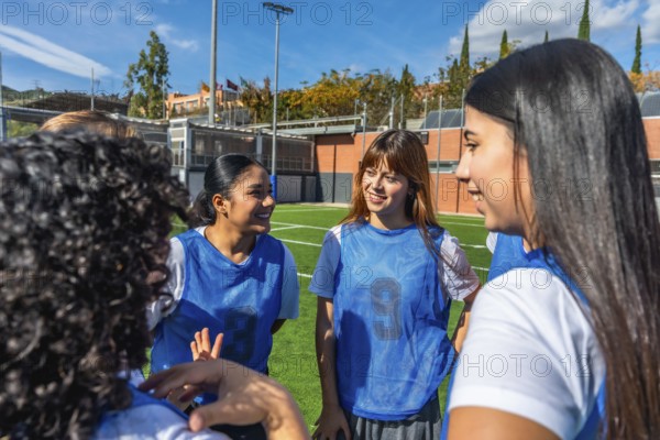 Female soccer teammates in blue bibs smiling and chatting on a sunny field, sharing a light moment during practice while preparing and bonding as a diverse, energetic team