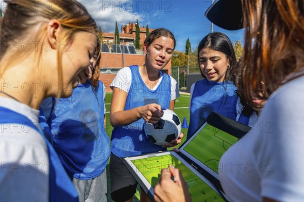 Women's soccer coach and team players discussing game strategy on a clipboard with a soccer ball, focusing on planning and teamwork on a sunny outdoor turf field