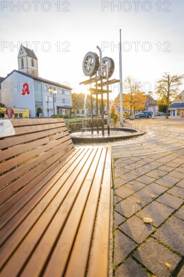 Municipal square with church and sculpture at sunset, concrete paving and autumn trees, Aidlingen, Böblingen district, Germany