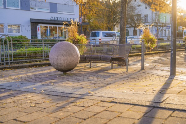 Public bench and large sphere on paved square, autumn trees and buildings in the background, Aidlingen, Böblingen district, Germany