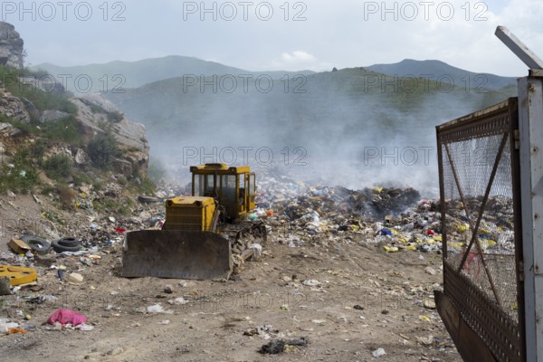 An excavator on a smoking garbage dump in the middle of mountains under a cloudy sky, Syunik province, Armenia