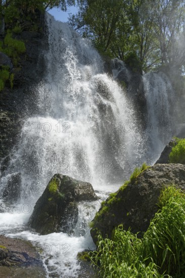 A waterfall falls over rocks surrounded by green vegetation and trees in sunshine, Shaki Waterfall, Syunik Province, Syunik, Armenia