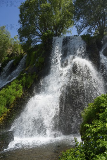 A waterfall flows over rocks surrounded by trees and green foliage under a blue sky, Shaki Waterfall, Syunik Province, Syunik, Armenia