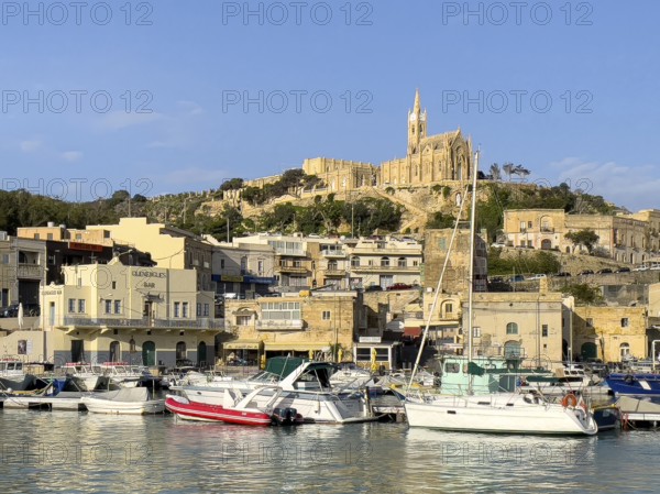 View of front sports boats sailboat in Mgarr harbour behind houses, on hills in the background parish church of Mgarr, Gozo, Malta
