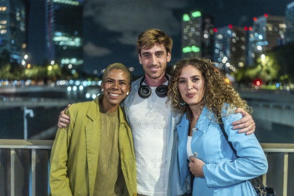 Diverse group of three friends standing arm in arm, smiling at the camera during a casual night out in an urban setting with city lights and blurred buildings in the background