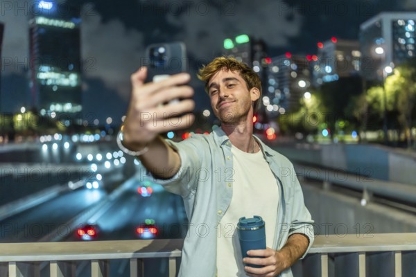 Young man smiling and taking a selfie with a smartphone, holding a travel mug on a bridge overlooking a busy highway with city lights illuminating the background at night