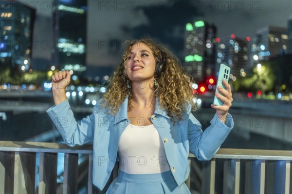 Young woman with curly hair wearing headphones and holding a smartphone, actively dancing to music on a city bridge at night, feeling happiness and freedom