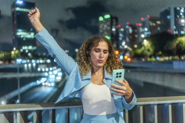 Young woman with curly hair wearing headphones and an elegant blue outfit, expressing joy and freedom while dancing to music on her smartphone at an urban overpass at night