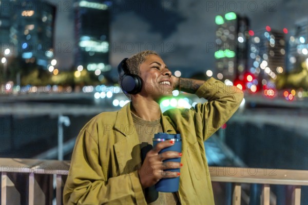 Happy black woman relaxing in a vibrant city at night, smiling while listening to music on wireless headphones and holding a reusable cup amid glowing urban lights and bokeh