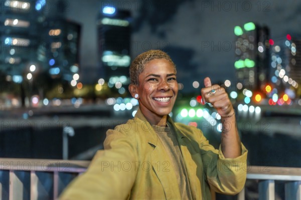 Happy young woman with short blonde hair and a radiant smile is taking a selfie gesture and showing a thumbs up, enjoying a vibrant urban night scene with blurred city lights in the background