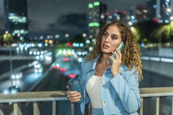 Professional businesswoman standing on a city bridge at night, having an important phone call with a reusable coffee cup in her hand, with urban traffic lights blurring in the background