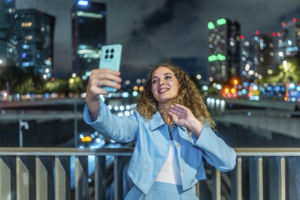 Young woman with curly hair and blue suit taking a selfie with her smartphone on a city bridge at night, bokeh lights from buildings and traffic visible in background