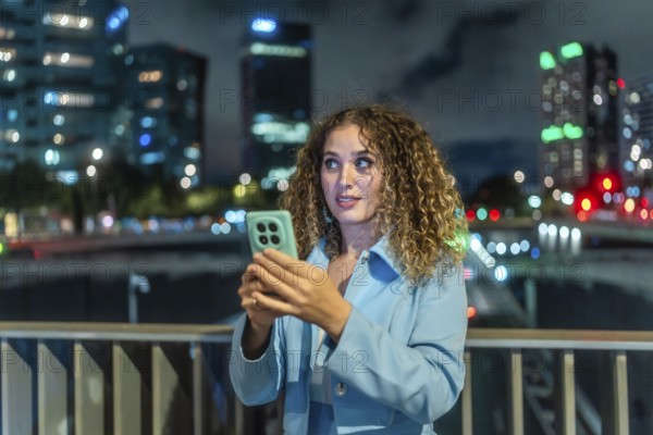 Young woman with curly hair looking surprised while holding a mobile phone, discovering new content or an interesting message in the busy city at night