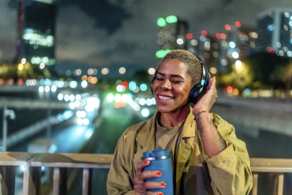 Woman standing on a bridge, holding a travel mug and wearing headphones while happily listening to music alone at night, with blurred city lights in the background