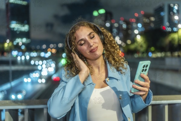 Young woman with curly hair finding joy and relaxation listening to music using headphones and a smartphone, standing on a bridge overlooking city lights and traffic at night