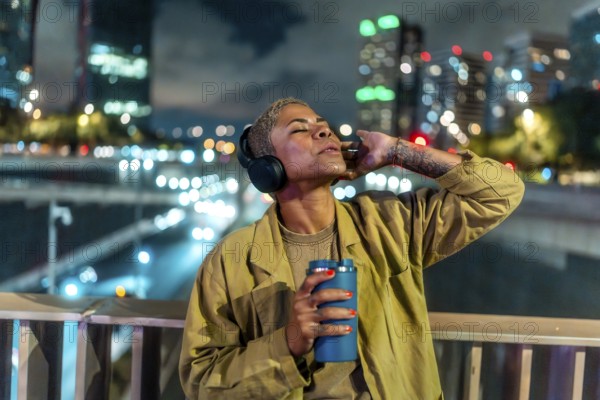 Woman standing on an overpass bridge in a city at night. Wearing headphones and holding a beverage container. With eyes closed. Relaxing and listening to music while city lights blur in the background