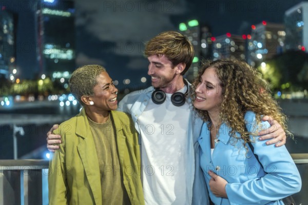 Three smiling diverse friends standing close together, enjoying a conversation and urban nightlife outdoors in the city with glowing building lights in the background