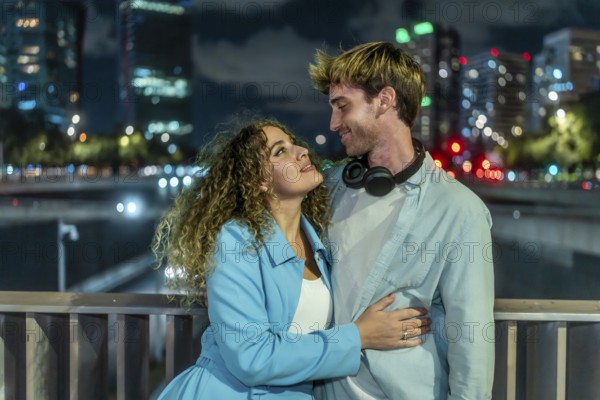Young couple embracing and looking into each other's eyes, expressing love and togetherness on a bridge overlooking a modern city skyline with blurred lights at night