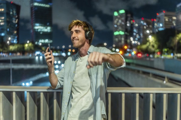 Young man with headphones and smartphone dancing joyfully on an urban bridge at night, blurred city lights bokeh creating a relaxed, modern nightlife vibe of freedom and fun