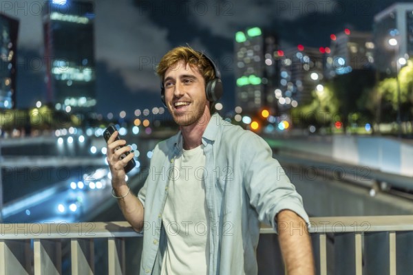 Smiling young man wearing headphones and holding a smartphone, happily listening to music while standing on a bridge in an illuminated urban city at night