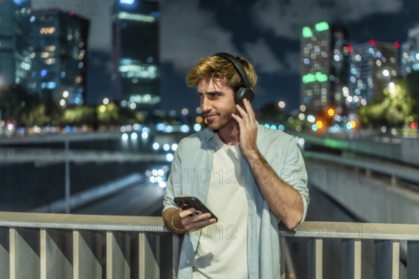 Young man is standing on a bridge, listening to music through headphones and holding a smartphone, with blurred city lights and traffic creating a vibrant bokeh background at night