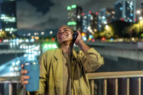 Young happy woman enjoying music through her headphones while standing on an urban bridge at night, with city lights and bokeh creating a vibrant background