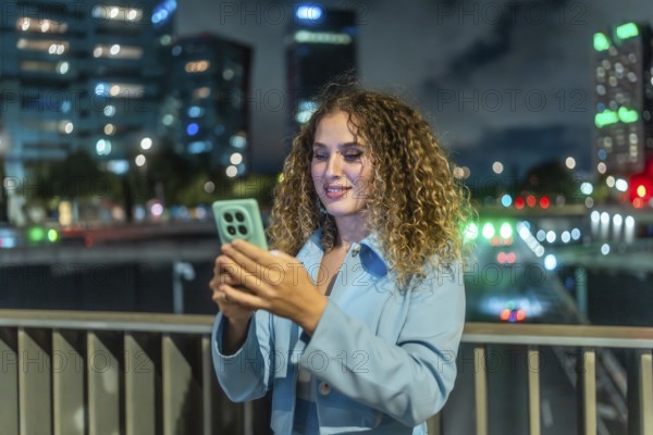 Young woman smiling, scrolling on her smartphone, enjoying connectivity and social media on a city street at night with blurry vibrant lights forming a bokeh background